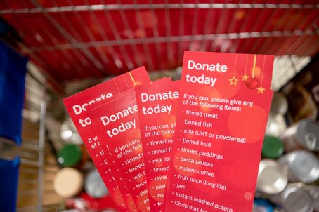 Close-up of red festive donation leaflets in a shopping trolley, listing items such as tinned meat, tinned fish, milk, fruit, and pasta sauces, encouraging people to donate food.