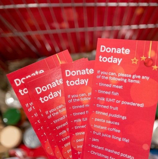 Close-up of red festive donation leaflets in a shopping trolley, listing items such as tinned meat, tinned fish, milk, fruit, and pasta sauces, encouraging people to donate food.