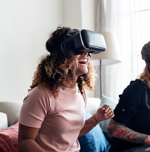 Two women laughing and enjoying a virtual reality experience while wearing VR headsets in a bright living room.