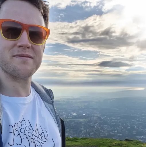 Jake stands on the Malvern Hills wearing orange sunglasses and a white T-shirt that reads “good good good good double good”, with a scenic view stretching out behind him under a dramatic cloudy sky.